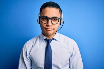 Young brazilian call center agent man wearing glasses and tie working using headset with serious expression on face. Simple and natural looking at the camera.