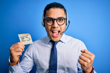 Young brazilian call center agent man holding reminder paper with yes message screaming proud and...