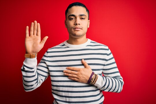 Young brazilian man wearing casual striped t-shirt standing over isolated red background Swearing with hand on chest and open palm, making a loyalty promise oath