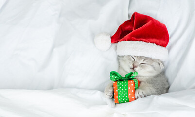 Funny kitten wearing red santa's hat sleeps under blanket with gift box. Top down voiew. Empty...