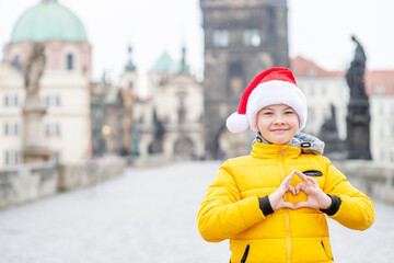 Happy young boy wearing red santa's hat shows heart sign on Charles Bridge in the Czech Republic. Empty space for text
