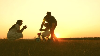 Mom and dad teach a young child to ride a bike at sunset in the park. Teamwork. Silhouette family walk
