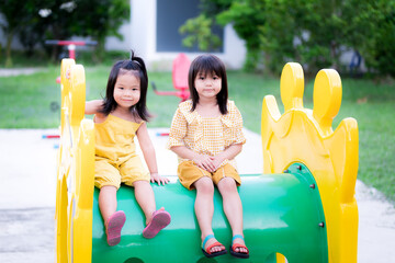 Obraz premium The two little girls smiled a little, sitting on a tube of yellow-green players. An Asian girl is wearing a yellow dress. Child is 3 years old and 5 years old.