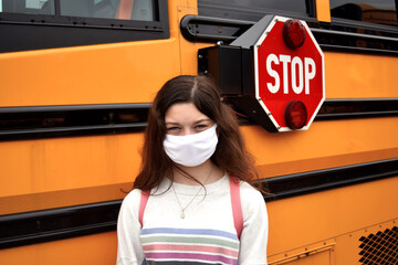 A female student, girl wears a face mask and backpack in front of a school bus. Coronavirus school reopening concept.