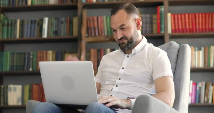 Mature man in white shirt is talking making video call, chat sitting in his office on bookshelves background. Remote work of teacher, professor or entrepreneur online at the computer, laptop.