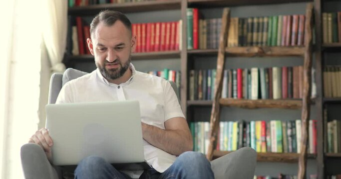 Mature man in white shirt is talking making video call, chat sitting on chair in his office on bookshelves background. Remote work of teacher, professor or entrepreneur online at the computer, laptop.