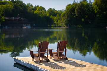 Two Adirondack chairs sitting on a wooden dock facing the calm water of a lake in Muskoka, Ontario...