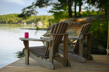 Two Adirondack chairs sitting on a cottage wooden dock facing the calm water of a lake in Muskoka, Ontario Canada. A purple insulated tumbler glass is visible on of of the chairs. - obrazy, fototapety, plakaty