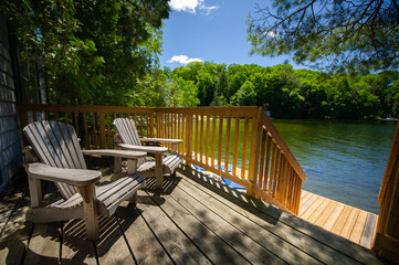 Adirondack chairs sitting on a cottage wooden deck facing a calm lake during a summer day in...