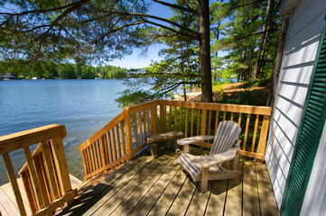 Adirondack chair sitting on a cottage wooden deck facing a calm lake during a summer day in...