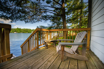Adirondack chair sitting on a cottage wooden deck facing a calm lake during a summer day in...