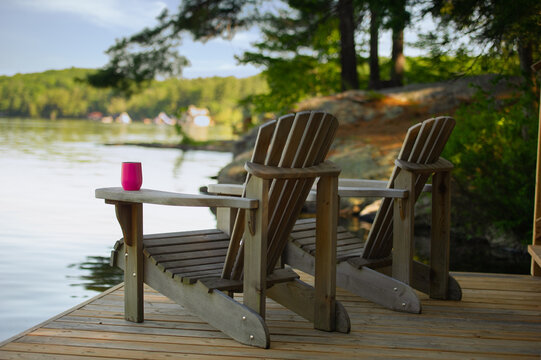 Two Adirondack chairs sitting on a cottage wooden dock facing the calm water of a lake in Muskoka, Ontario Canada. A purple insulated tumbler glass is visible on of of the chairs.
