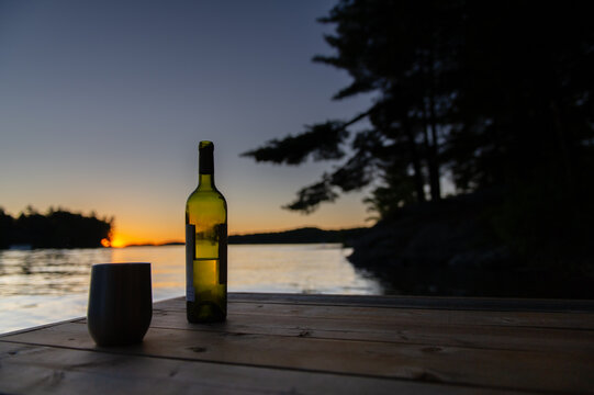 Bottle Of Wine And A Tumbler Glass On A Cottage Wooden Dock Facing The Calm Water Of A Lake During Sunset.