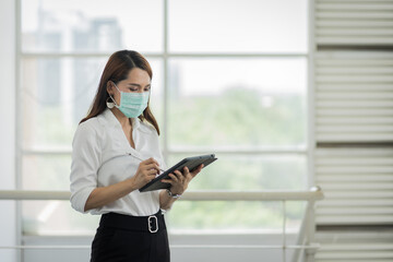 Fototapeta na wymiar Portrait of a young adult Asian business woman in white shirt and black long pants wears surgical facemask looking on tablet in company building during VOCID-19 pandemic. Business stock photo.
