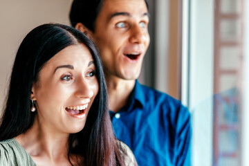 Wow. Close up portrait of young couple, man and woman looking excited through window together, while standing indoors. Focus on dark haired woman. Horizontal shot