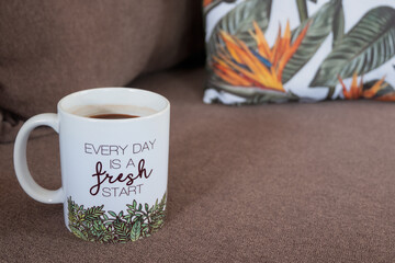 Coffee mug with inspirational message on couch with throw pillows. Every day is a fresh start. Copy space.