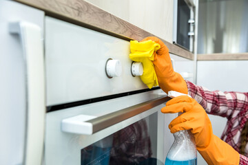 Young wife cleaning oven with yellow rag and spray in kitchen