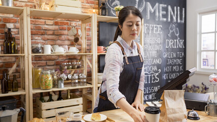 smiling young asian girl bartender standing in wooden counter in vintage cafe store and prepared ready for customer order. barista lady putting takeaway paper bag and cup on table in coffeehouse.