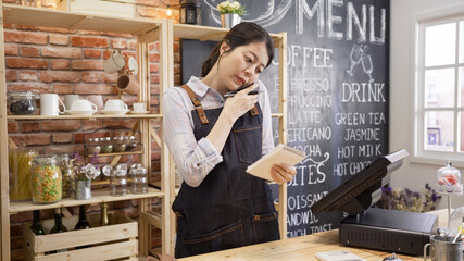 small business communication people and service concept. waitress in denim apron calling on smartphone at bar counter in coffee shop. coffeehouse staff holding note talk with customer taking order