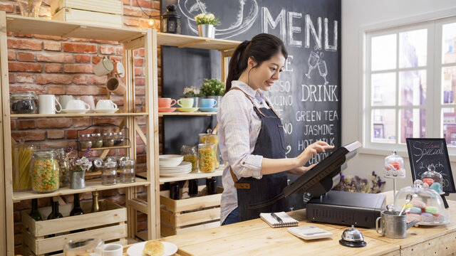 smiling coffee shop assistant using pos point of sale terminal to put in order from note paper at restaurant register. waitress lady working in counter in cafe store. young girl staff using cashbox.