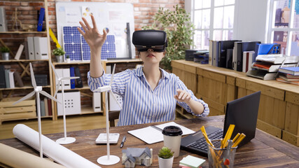 architect in vr headset at workplace with laptop schemes and building models in office. young girl engineer worker wearing virtual reality goggles sitting working on green energy construction plan