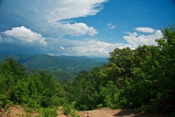 A country road runs along the side of the mountain. The forest is around. A large thundercloud appears in the blue sky over the mountains. Summer mountain landscape.