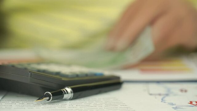 blurry man finger counts currency on black outdated digital calculator sitting at table with pen on papers slow motion closeup