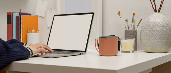 Female designer working with mock up laptop on white table with painting tools, books and decorations