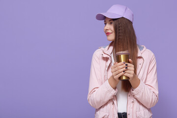 Portrait of thoughtful woman wearing baseball cap, drinks coffee from thermo mag, has deep thoughts during break, adores favorite drink, poses against lilac wall, looking aside.