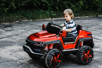 curly-haired boy in a striped T-shirt rides a red big toy car driving on an asphalt path. day off, outdoor recreation