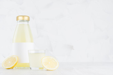 Fresh yellow lemon juice in glass bottle mock up with blank label,  wine glass, fruit slice on white wood table in light interior, template for packaging, advertising, design, branding product.