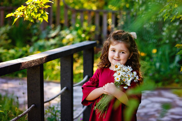 Fototapeta premium Summer portrait of a charming little girl with a bouquet of daisies in nature. A girl in a red dress holds a bouquet, smiles and looks at the camera.