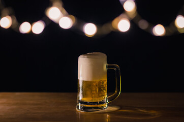 A glass of beer on wooden table that have bokeh lights on top with dark background.