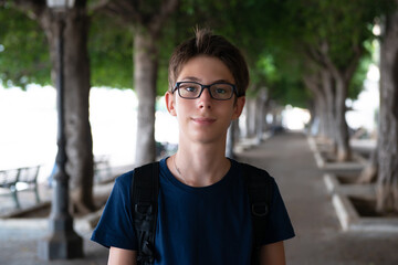 Young boy walking in summer park with trees near the sea. Cute spectacled smiling happy teen boy 13 years old, looking at camera outdoor. Kid's outdoor portrait.