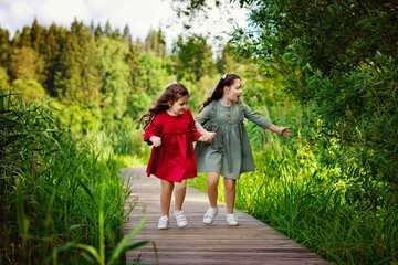 Naklejka na ściany i meble Two little girls with long hair in a green and red dress run along a wooden path in the Park. A fun childhood of the two sisters.