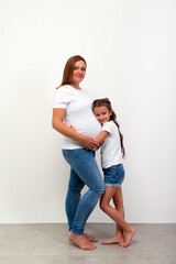 Pregnancy. Happy Family Expecting Baby. Beautiful smiling girl hugs her pregnant mother with big belly. Indoor portrait of mom and daughter.