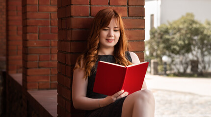 European female student with red hair sitting near the library and reading a book. Self-education concept