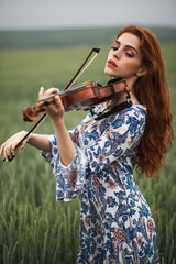 Beautiful romantic girl with red hair and blue dress holding violin on nature field of  flowers....
