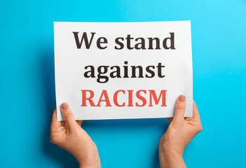 Female hands with text WE STAND AGAINST RACISM on paper, top view