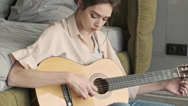 Young amazing concentrated woman indoors at home sitting near bed while playing on a guitar