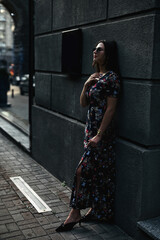 Midshot of amazing woman in sunglasses outdoor. Beautiful brunette poses on the street in contouring sunlight, she stands near wall and holds hands up near head, eyes closed. Copy space, vertical shot