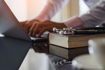 Male doctor hands typing on laptop computer keyboard, search medical information with text book and medical stethoscope on the desk at office. Online medical,medic tech, emr, ehr concept.