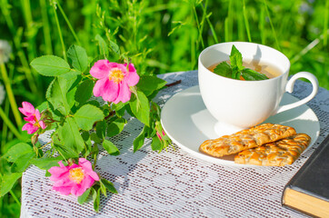 A Cup of green tea with a mint leaf, flowers, and a book on a table in the garden. Evening tea with cookies. Summer village concept. Copy space.