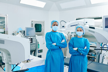 Obraz premium Confident young surgeon assisistants in scrubs and medical masks standing in operating room with arms crossed and looking at camera