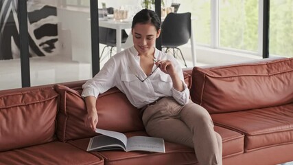 Smiling elegant pretty asian woman reading magazine while holding eyeglasses and sitting on sofa at home - Powered by Adobe
