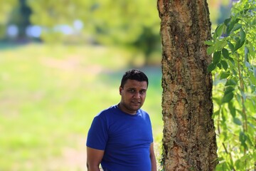 close portrait of young man in blue t shirt standing outside in park with trees in background