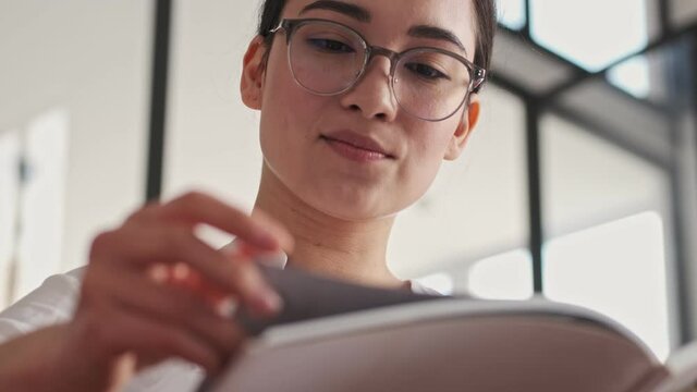 Pleased elegant pretty asian woman in eyeglasses reading magazine while sitting on sofa at home