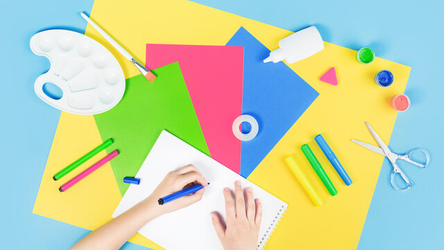 Hands drawing over color paper, scissors, pencils paint and various school stationery on blue yellow background. Flat lay with copy space for back to school or education and craft concept