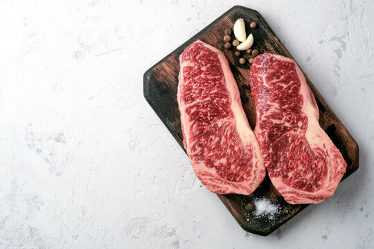 Two raw strip steaks on a chopping Board with seasonings and garlic on white stome, top view and copy space