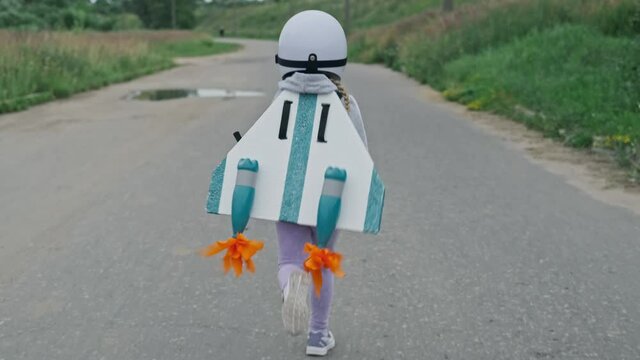 Little girl wearing white helmet, jetpack and running on rural road in slow motion. Tracking of kid pretending to be pilot. 4K, UHD